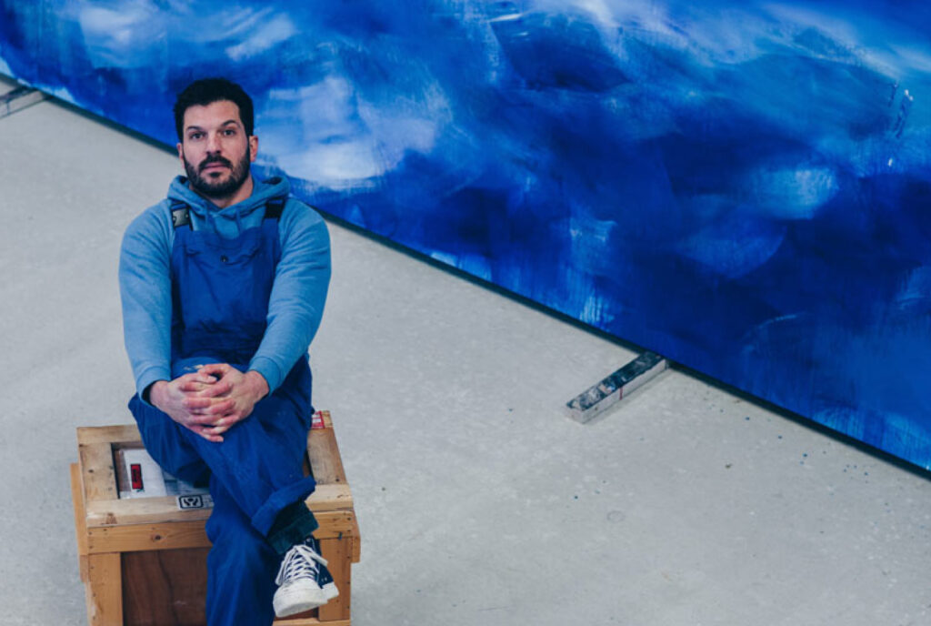 Artiste assis sur une caisse, devant une œuvre à la Salle d’exposition Martres Tolosane.